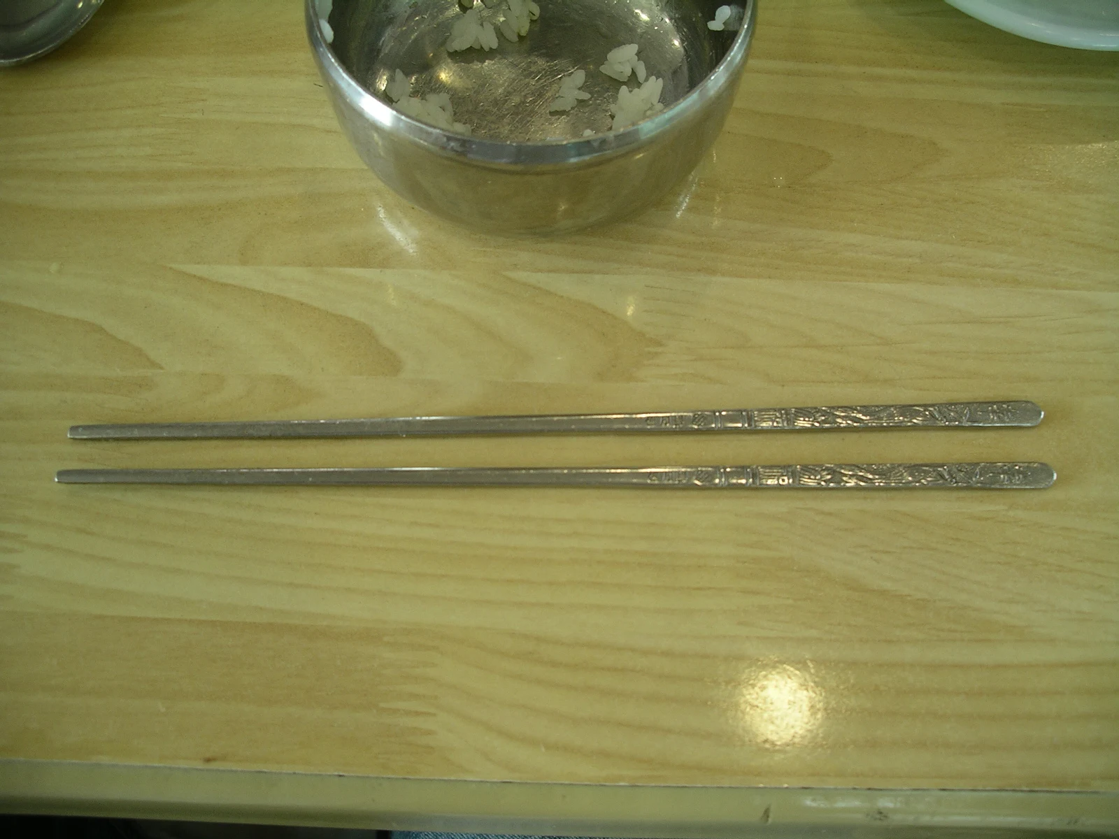 A pair of decorated Korean metal chopsticks resting beside a stainless steel rice bowl.