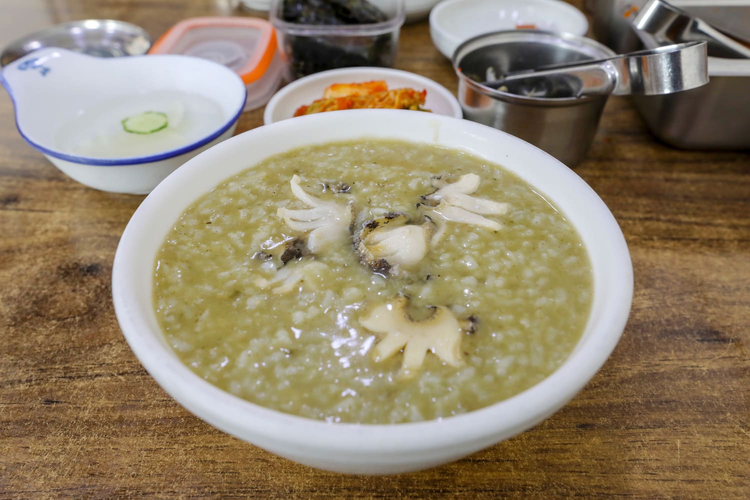 A white bowl of Korean abalone porridge, greenish from the abalone, with sliced abalone on top, served on a wooden table alongside kimchi, seaweed, water kimchi with a cucumber slice, and stainless steel dishes.