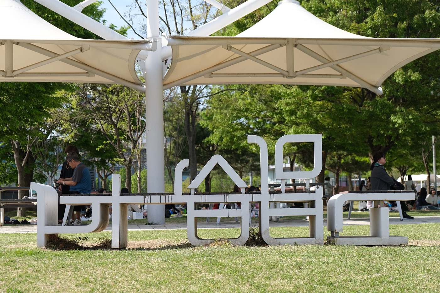 Letter-shaped benches spelling 뚝섬 under white canopies in Ttukseom Hangang Park, Seoul, in April 2026, with visitors on the lawn behind.