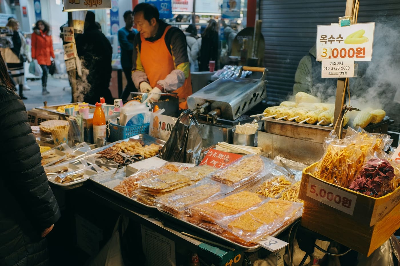 A Korean market street-food stall with Korean price signs, grilled corn skewers, and packaged snacks.