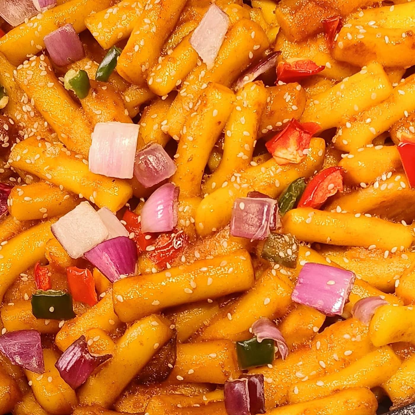 A close-up of tteokbokki rice cakes coated in sauce with sesame seeds, onion, and chili.