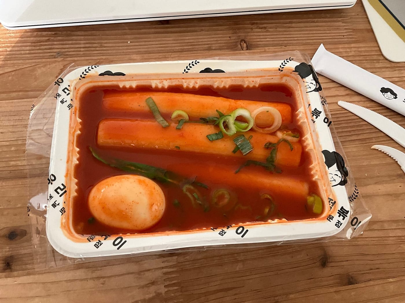 A takeout tray of tteokbokki with thick garaetteok rice cakes, a boiled egg, and green onion in red sauce.