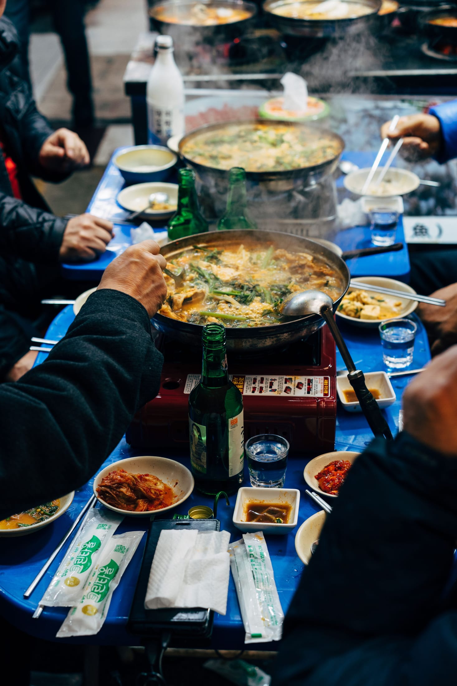 Friends and family sharing a Korean meal at a restaurant table with soju bottles, kimchi, and a shared hot pot.