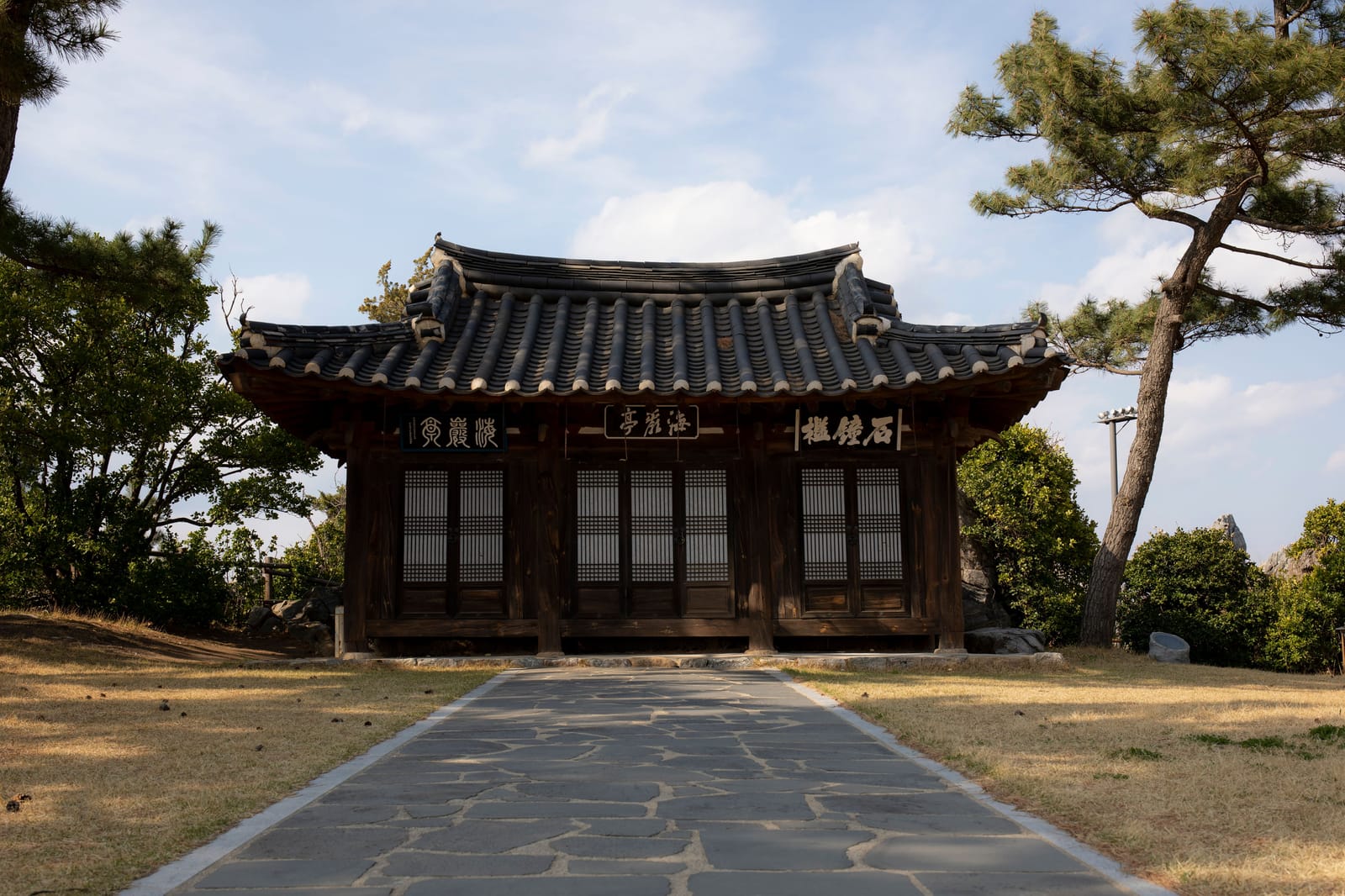 A traditional Korean wooden pavilion with a tiled roof, surrounded by pine trees under a clear sky.