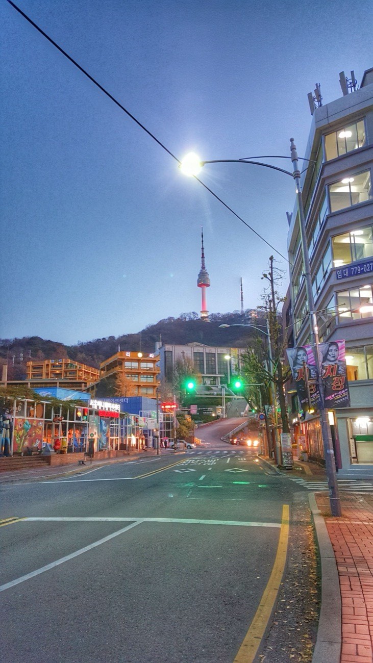 N Seoul Tower lit at dusk on top of Namsan, seen from a street at the foot of the mountain with traffic lights and street lamps on.