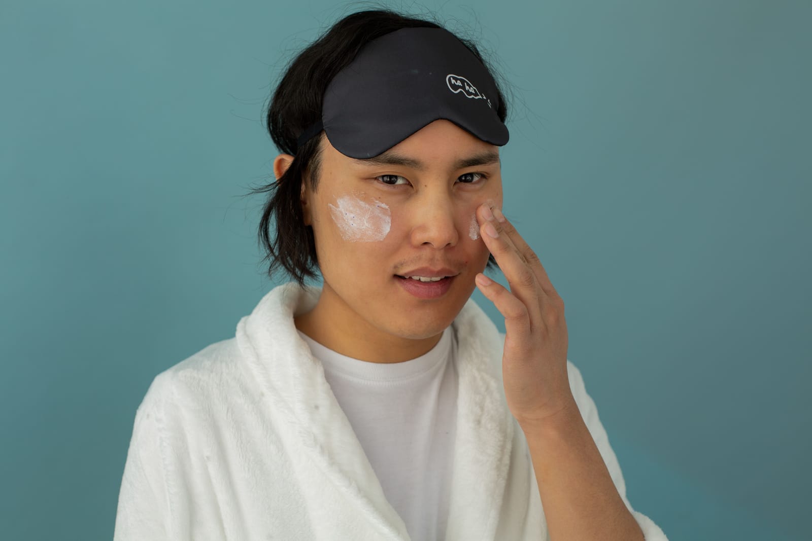 A man applying moisturizer to his cheek, wearing a bathrobe and sleep mask.
