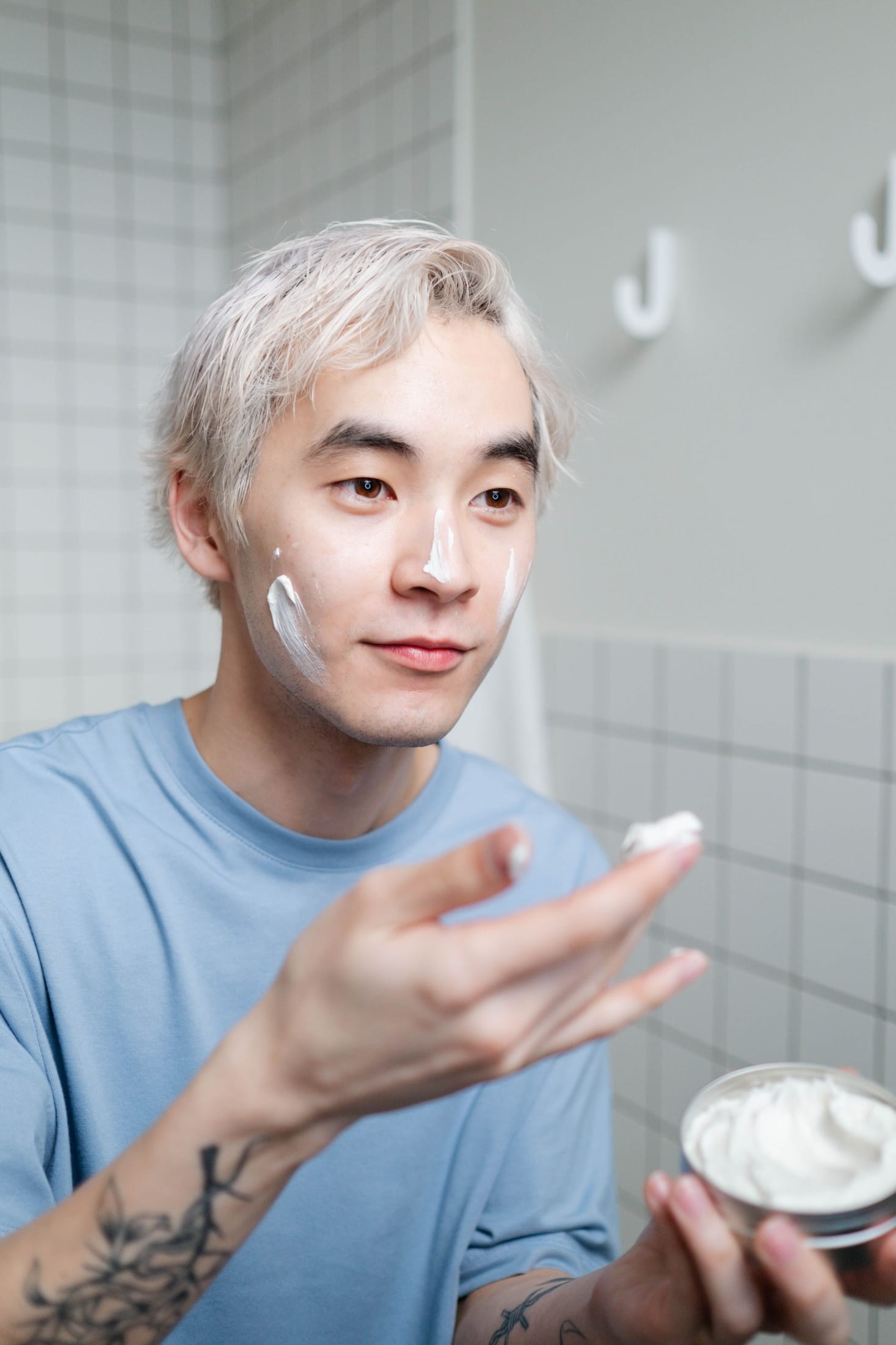 A man with light hair applying face cream in a tiled bathroom, holding an open jar.