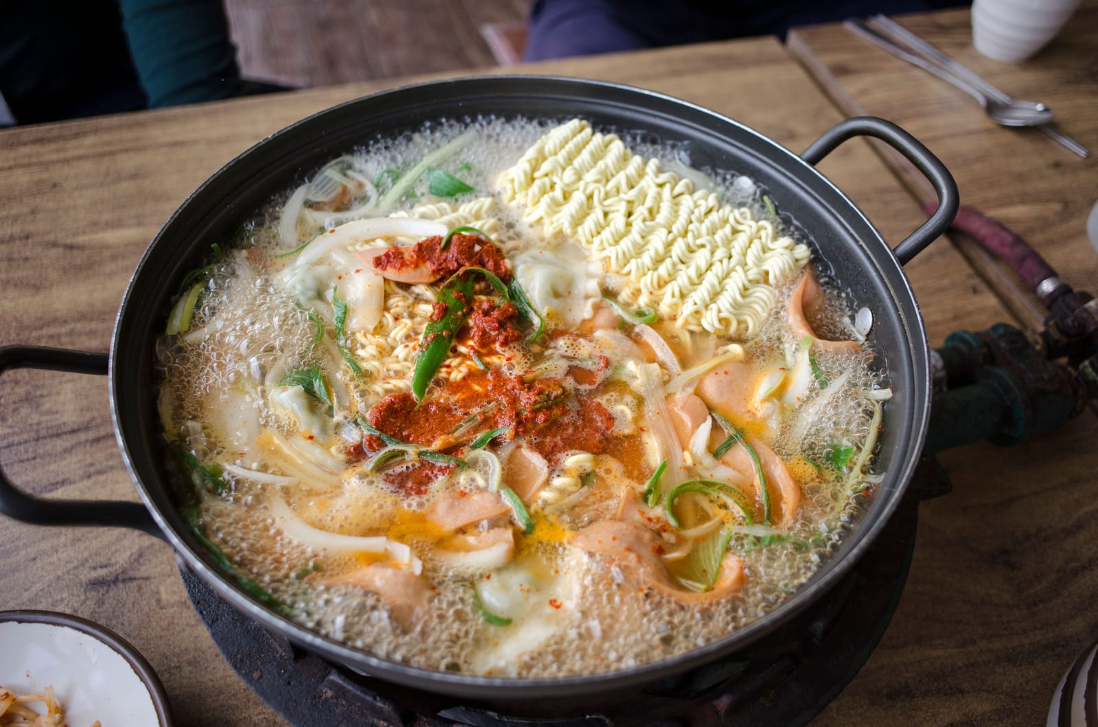 A bubbling pot of budae jjigae with sausage, dumplings, instant noodles and chili paste on a tabletop burner.