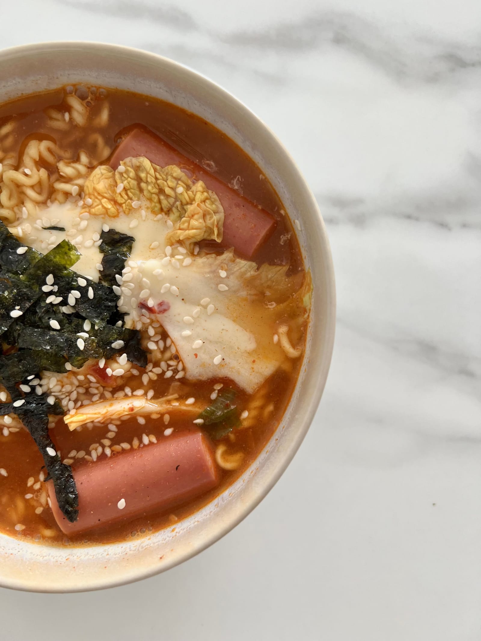 A bowl of spicy Korean instant noodles topped with Spam, seaweed, melted cheese and sesame seeds.