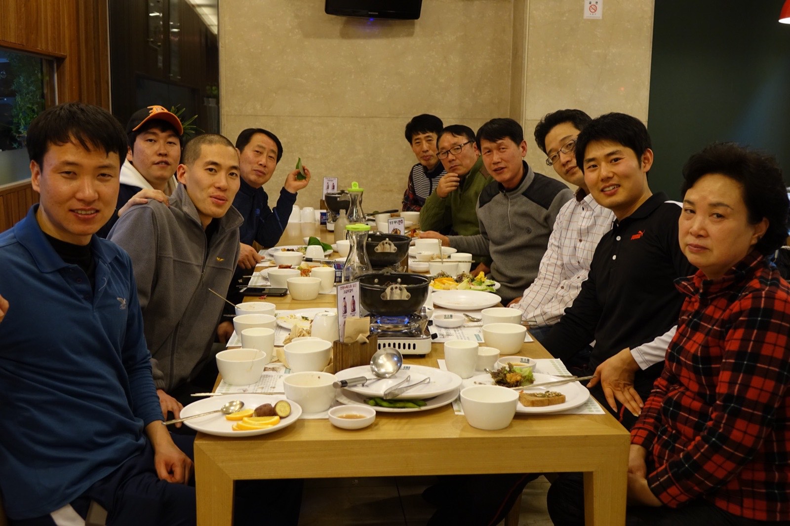 A team of Seoul fire-station staff seated at a long restaurant table with hotpots and side dishes during a team dinner.