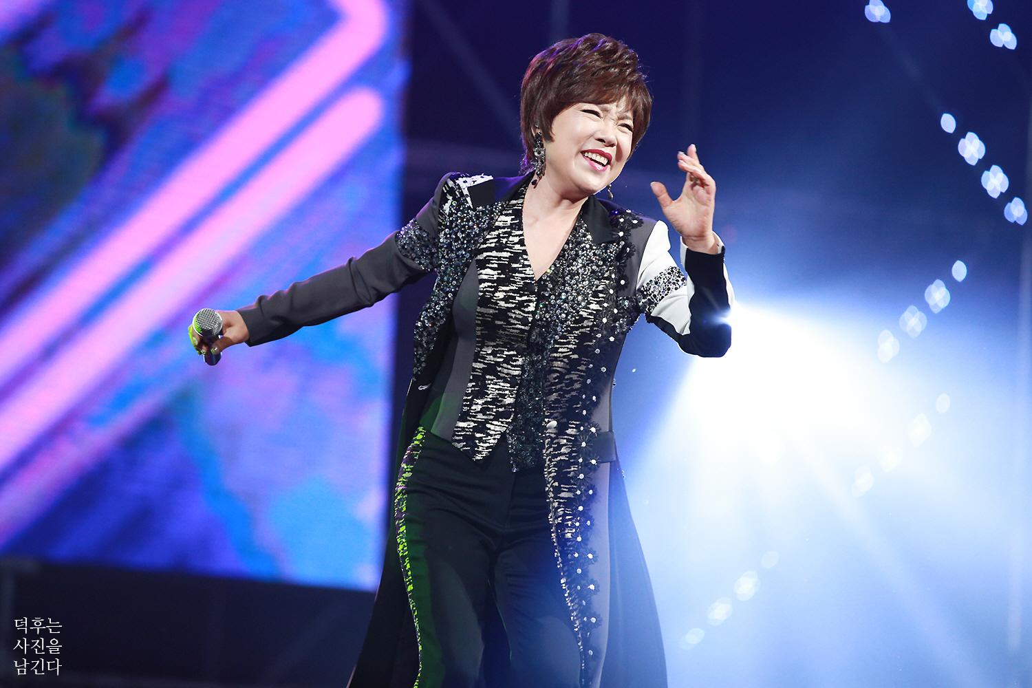 Trot singer Kim Yon-ja smiling and gesturing on stage with a microphone under blue concert lights.