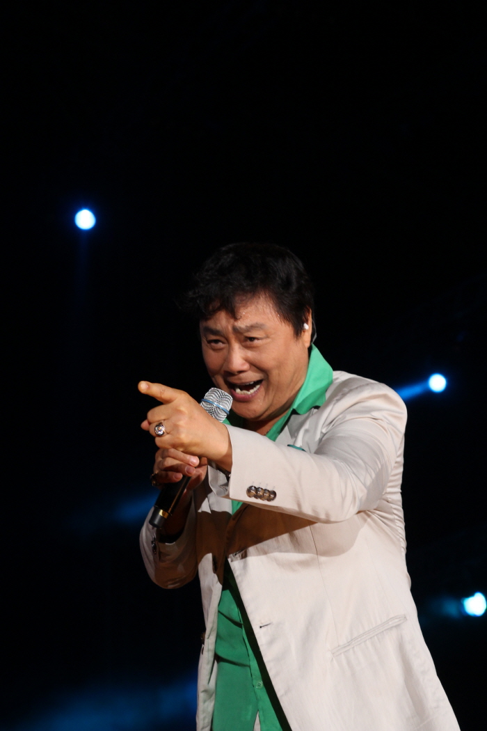 Trot legend Nam Jin performing on stage with a microphone, singing under blue stage lights.