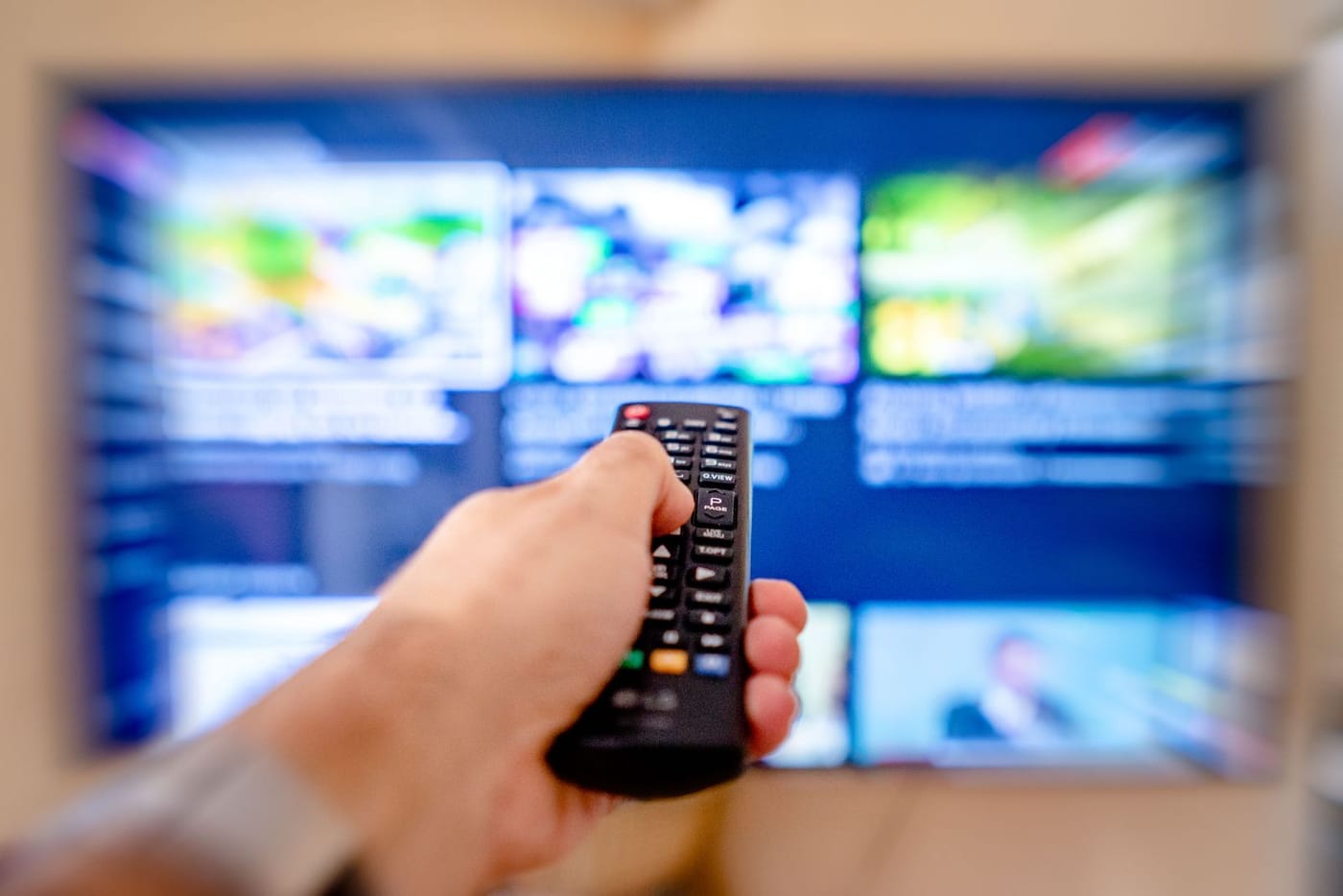 A person holding a TV remote in front of a screen showing a blurred, generic multi-thumbnail streaming layout.