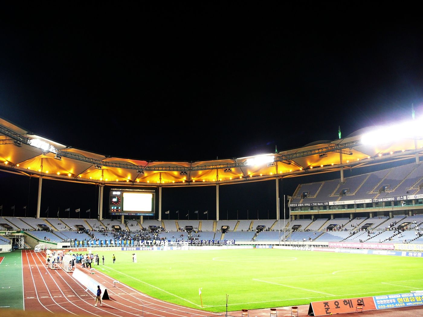 The main stadium at Incheon Munhak Sports Complex at night, its curved roof canopy lit amber over the stands, with players on the floodlit pitch and an athletics track.