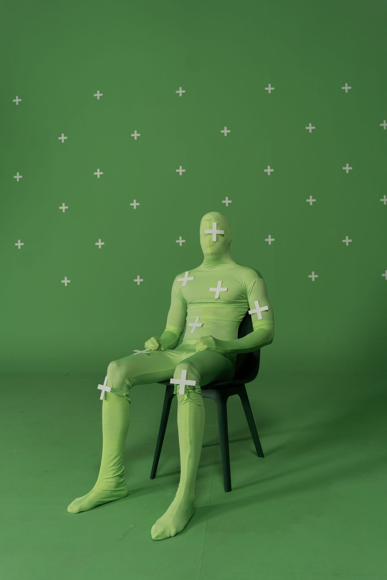A real person wearing a chroma-key motion-tracking bodysuit with tracking markers, seated in a studio setup.