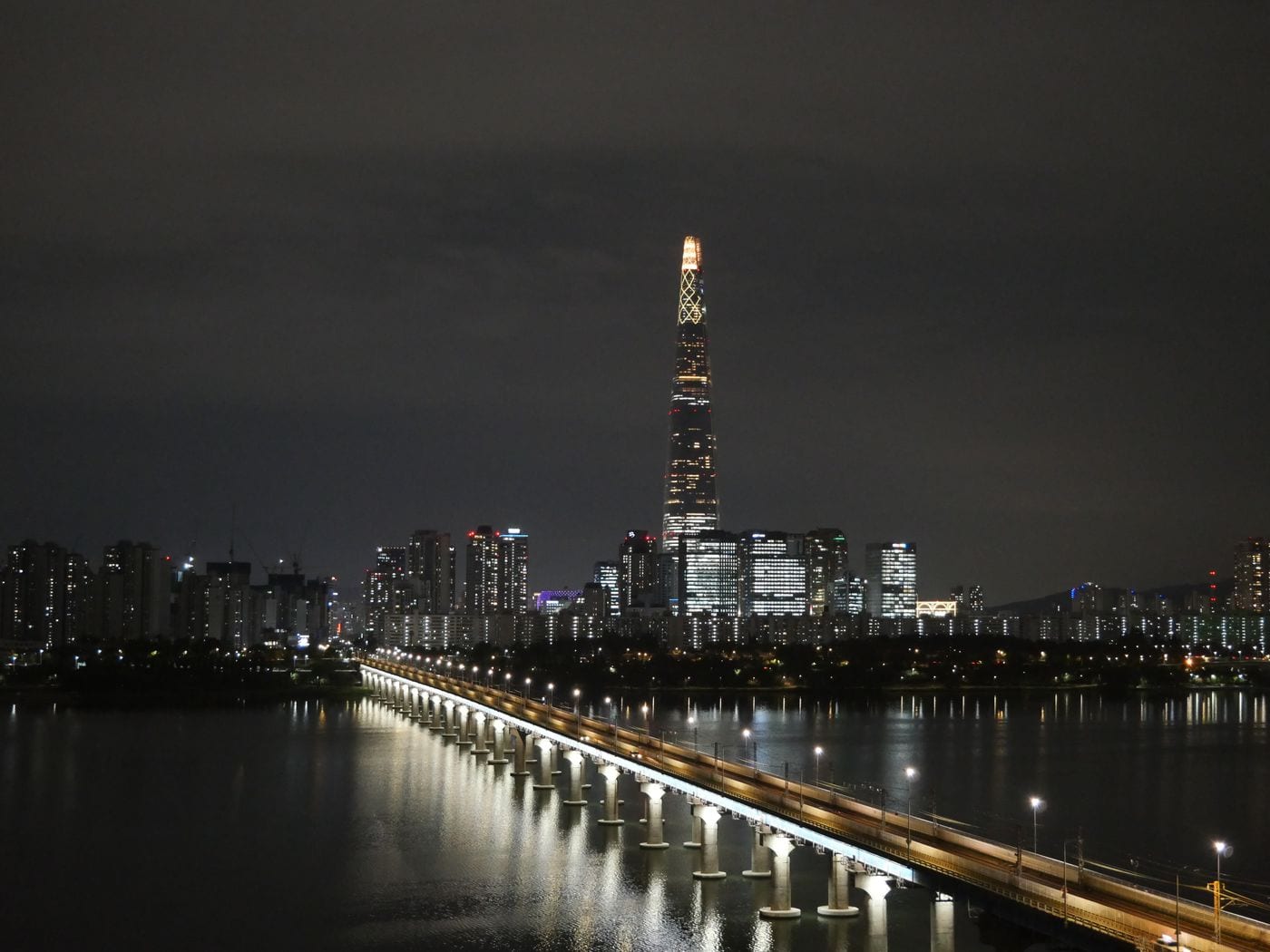 A night view of the Seoul skyline and Han River, with Lotte World Tower lit up and a river bridge in the foreground.