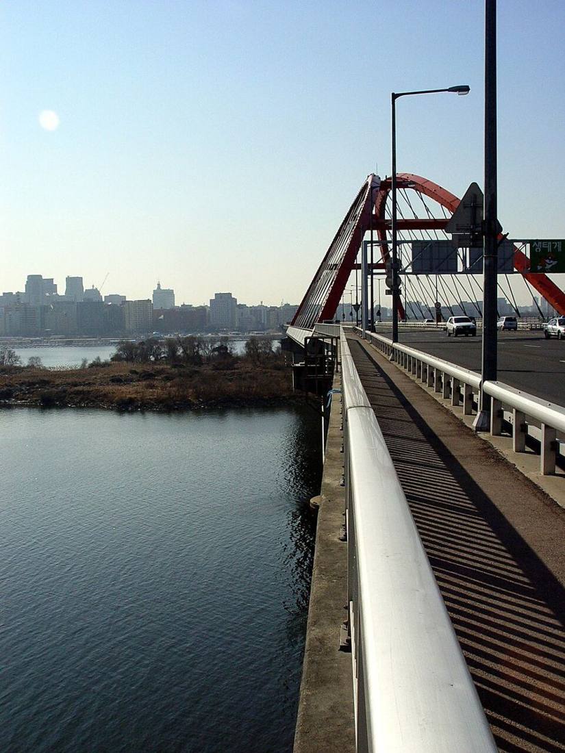 The red steel arch of Seogang Bridge in Seoul, seen from the bridge's sidewalk, with the Han River and Bamseom islet below and the skyline in the distance.