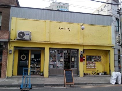 The front of Hanmi Bookstore in Incheon, a low building painted bright yellow with the name 한미서점 in black letters, bookshelves visible through the windows and a chalkboard sign on the pavement.