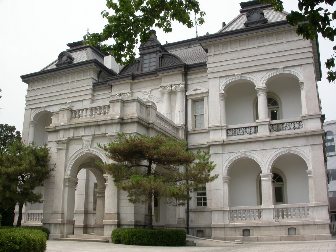 The Western House of Unhyeongung, a pale stone two-storey building with arched balconies on both floors, columns and a mansard roof, with a pine tree in front.