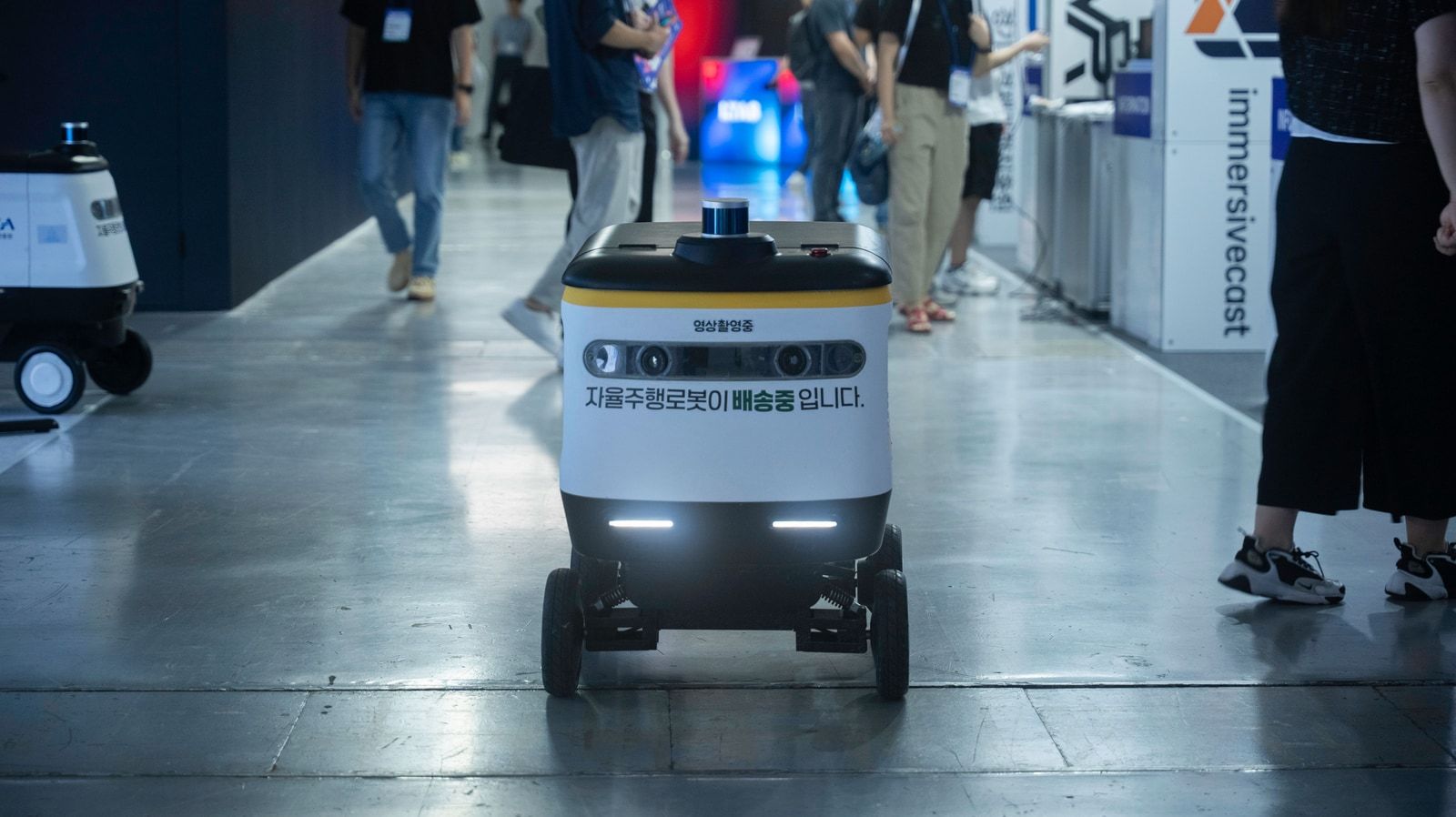 A real autonomous delivery robot navigating an aisle at a Korean technology conference, with Korean-language signage visible on its body.