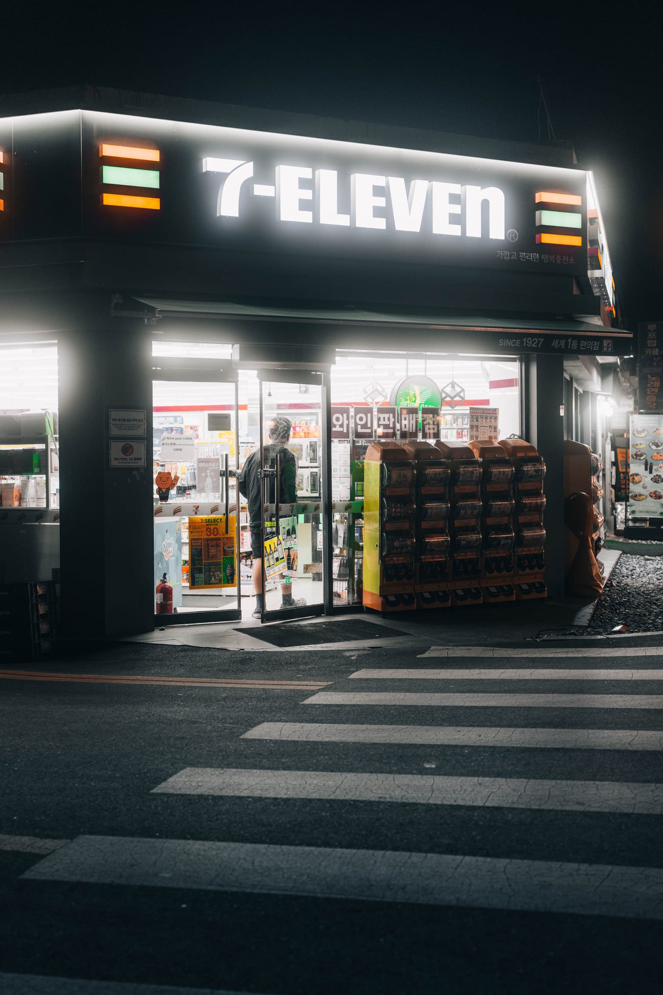 A 7-Eleven in Seoul at night, lit up on a corner with Korean signage.