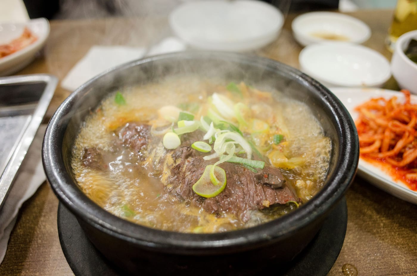 A steaming Korean beef soup in a black stone pot, topped with sliced spring onion, with kimchi alongside.