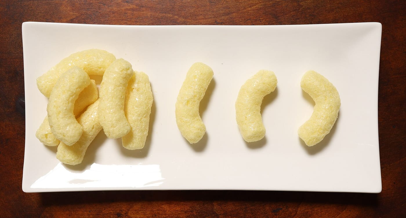 Five pale corn puffs on a white rectangular plate — each curved like a small banana, with a lightly pitted, porous surface.
