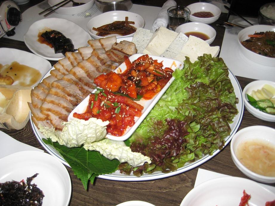 A Korean restaurant bossam platter: sliced boiled pork belly, red bossam kimchi, tofu, napa cabbage leaves, lettuce and perilla leaves, with a bowl of salted shrimp beside it.