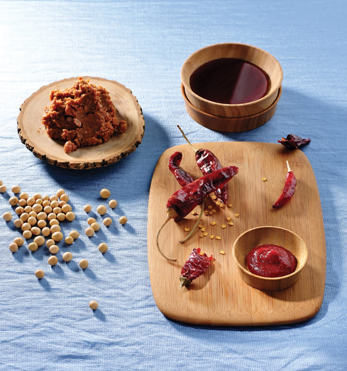 Soybeans, a mound of brown doenjang, a bowl of dark soy sauce, dried red chillies and a small bowl of gochujang arranged on a blue cloth.