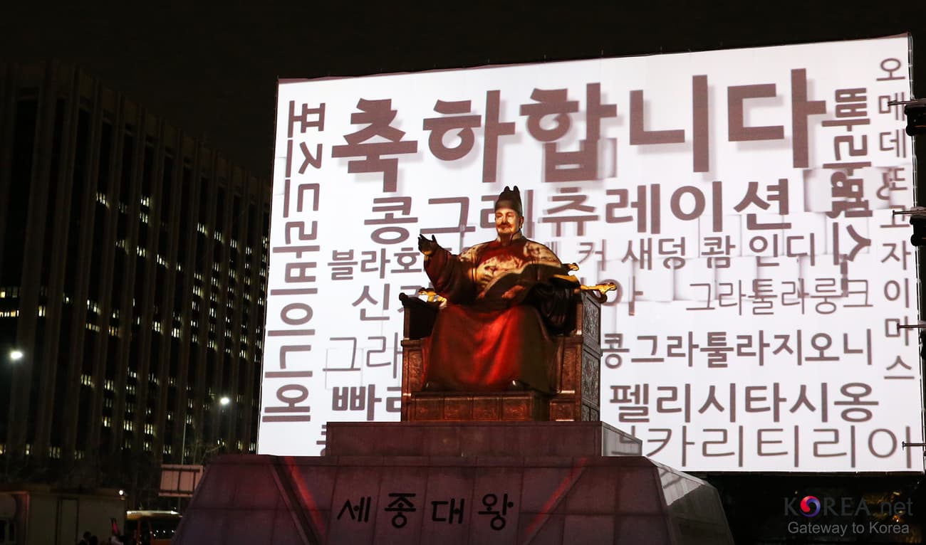 A night projection on the Sejong Center facade reading 축하합니다 in large Hangul, surrounded by the word "congratulations" written phonetically in Hangul in many languages, with the Sejong statue silhouetted in front.