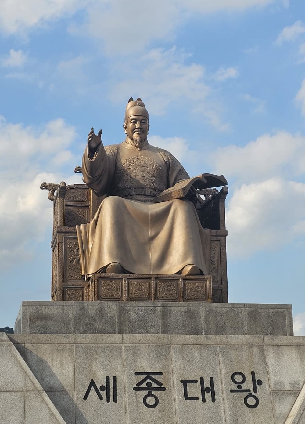 Close view of the bronze King Sejong statue at Gwanghwamun against a blue sky, seated with an open book, the granite pedestal reading 세종대왕 in Hangul.
