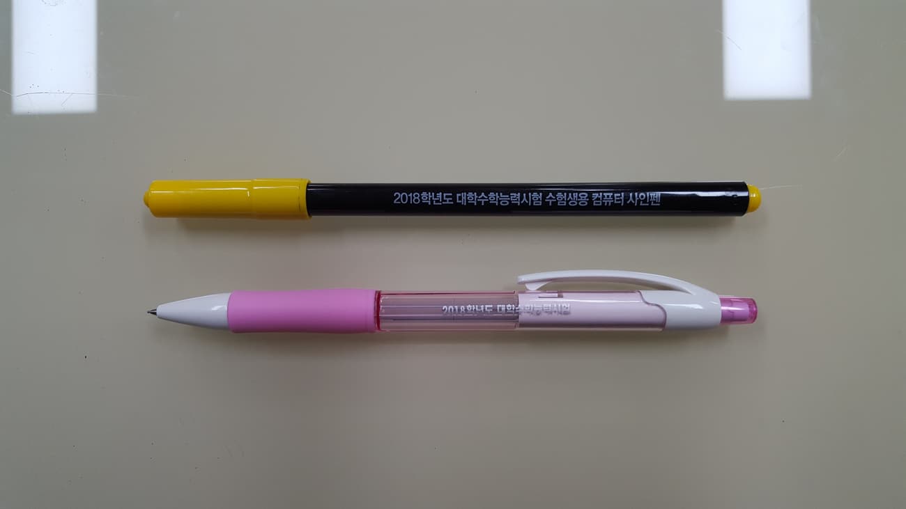 A black computer-use marker pen and a mechanical pencil laid on a white surface — the two writing tools required for the Korean exam.