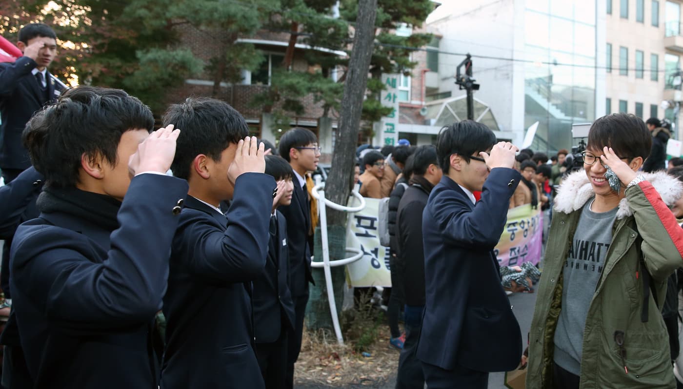 Uniformed junior high-school students lined up along a road, saluting with hands raised to their foreheads as older students walk past to the exam hall.