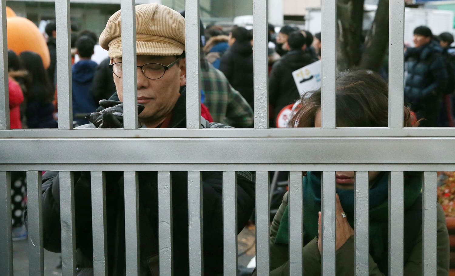 Parents pressed against the metal railings of a school gate on Suneung morning, one man with his eyes closed and hands raised in prayer, a crowd of people behind him.