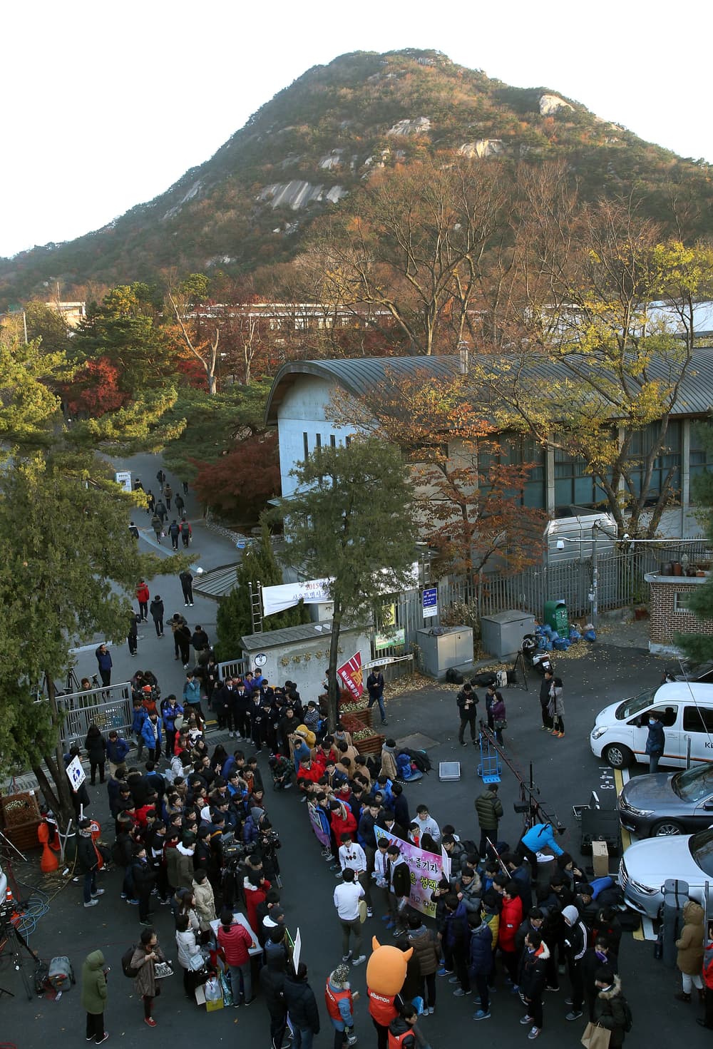 Aerial view of a Seoul high school gate on exam morning: a dense crowd of students, parents, banners and TV crews on the road below, with an autumn-coloured mountain rising behind the school.
