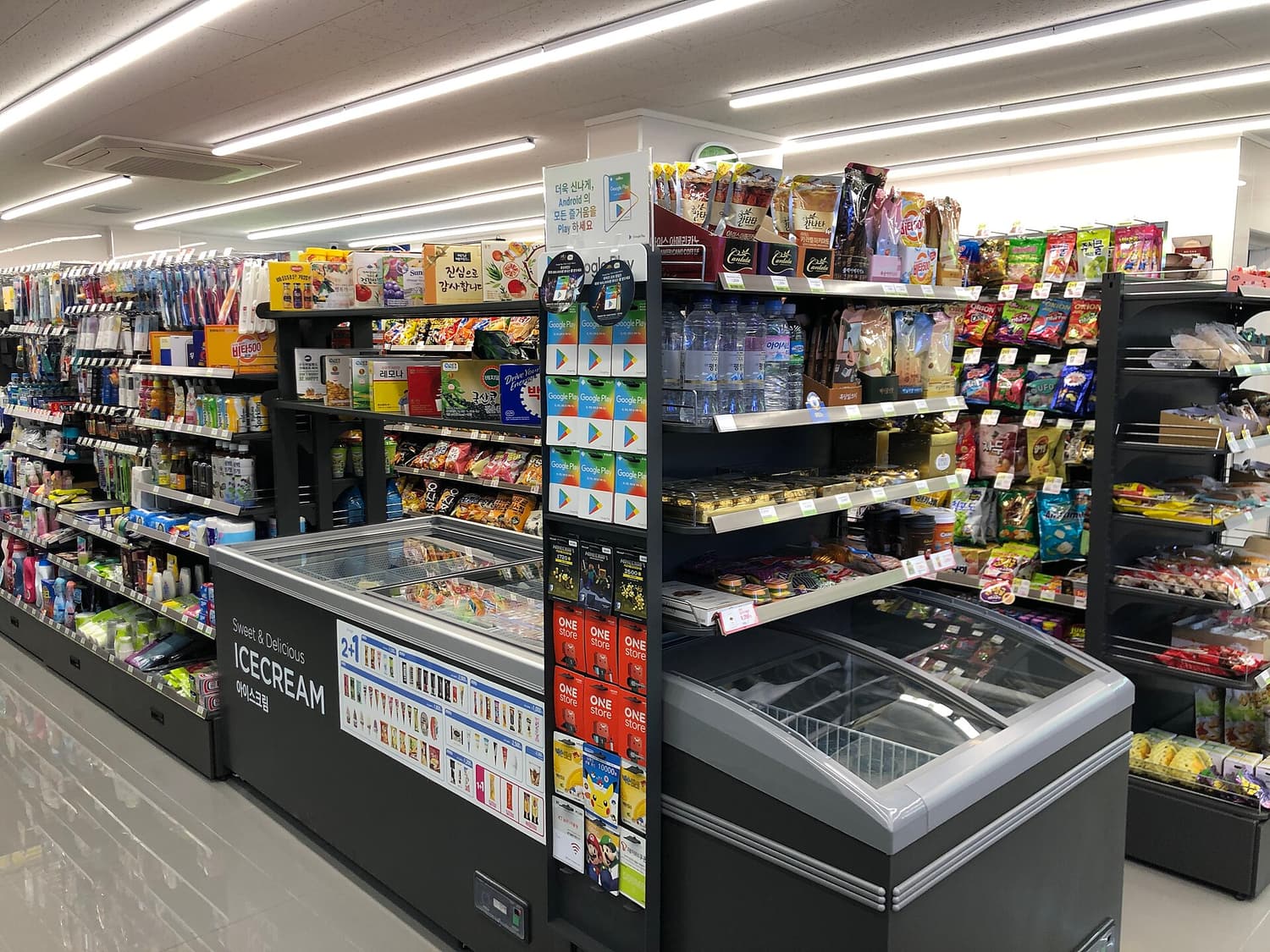 Interior of a CU convenience store in Korea. The ice-cream chest in the centre carries a 2+1 promotion strip showing a grid of eligible products, with snack shelves, a drinks wall and gift-card racks behind it.