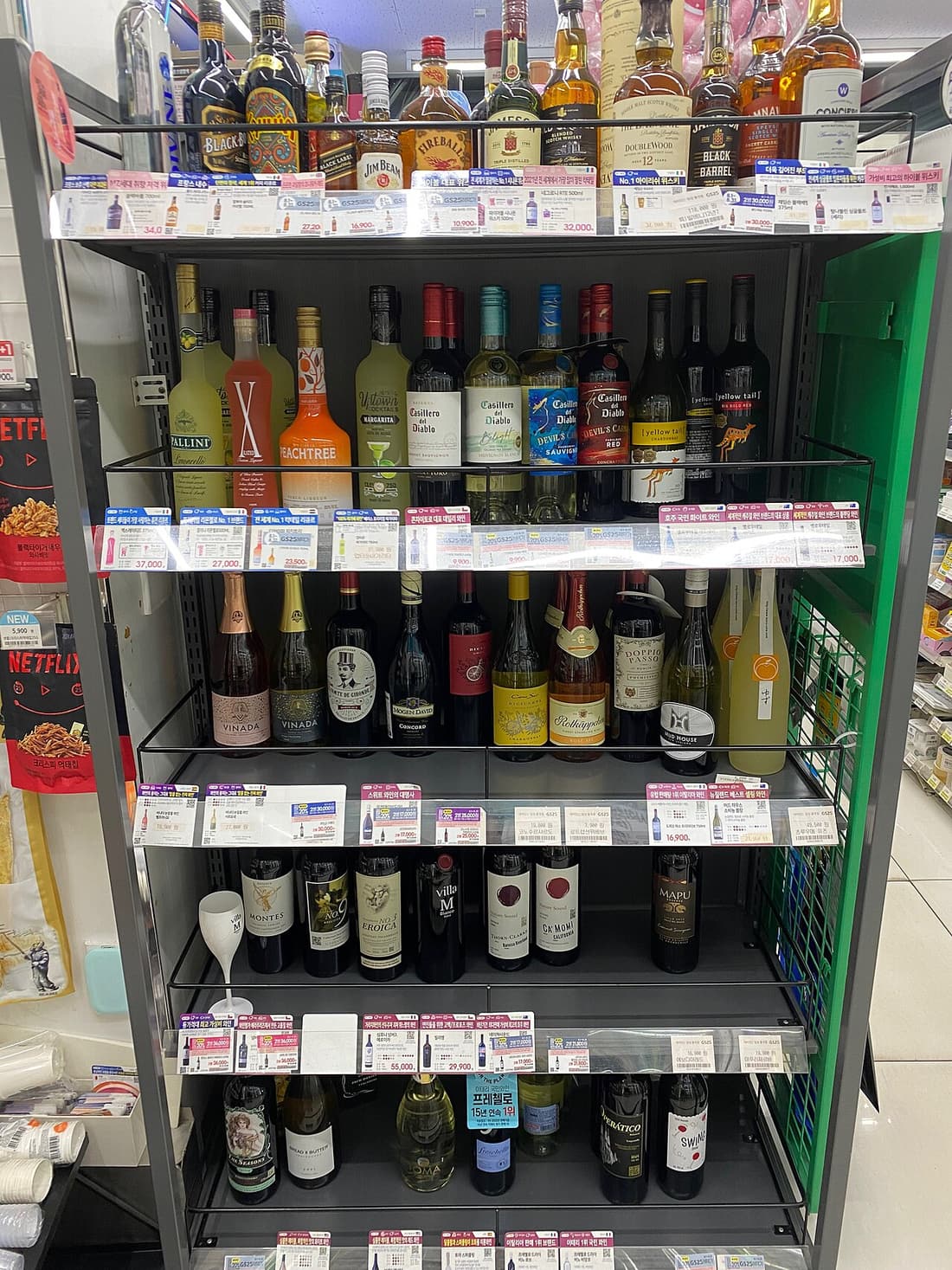 A GS25 shelf in Seoul photographed straight on, with a dense row of printed price tags clipped along the front edge of every shelf, several of them carrying coloured GS25 promotion flashes.