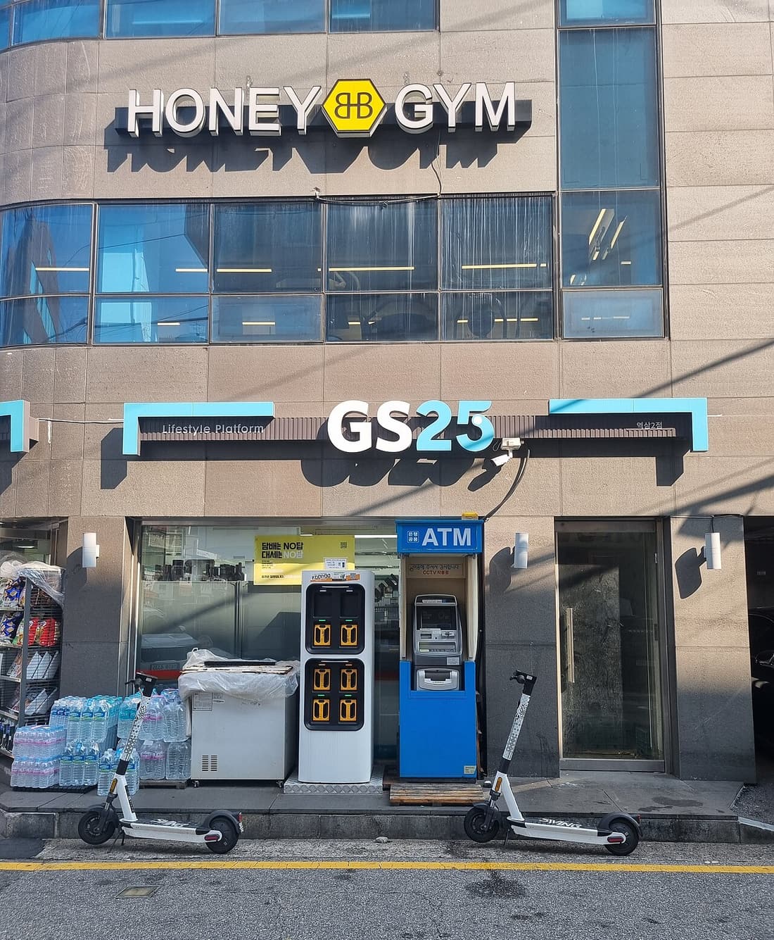 Street-level view of a GS25 branch in Yeoksam, Seoul, with the store sign above the window, a bank ATM beside the door, an electric-scooter battery swap cabinet and stacked cases of bottled water on the pavement.