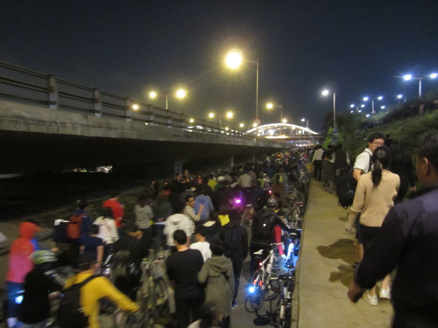 A dense crowd of people and cyclists filling a narrow riverside path under a bridge at night after the fireworks festival, moving slowly in one direction.