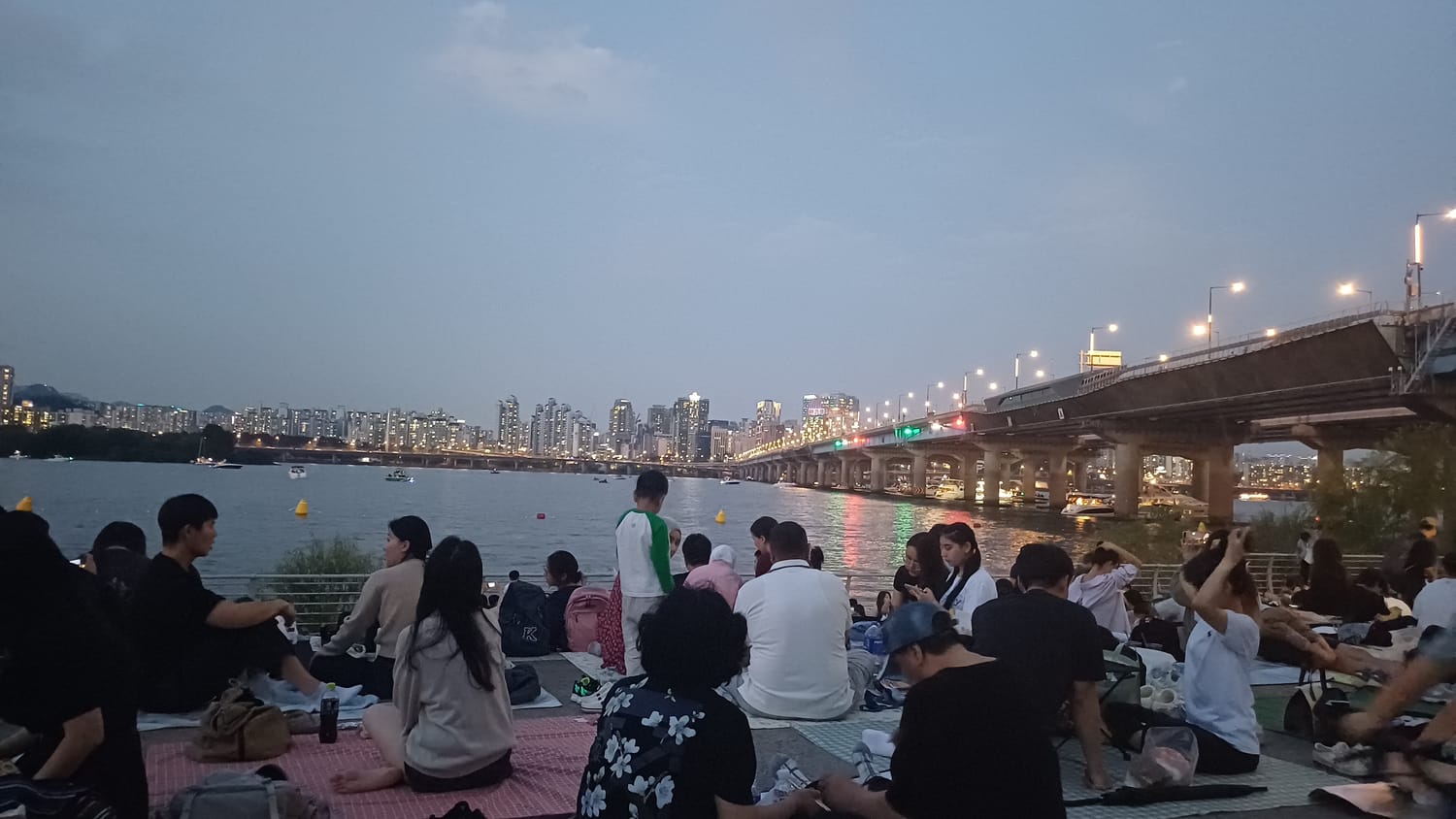 Crowds sitting on picnic mats along the Yeouido riverbank at dusk, with the Han River, small boats, Mapo Bridge and the Seoul skyline behind them.