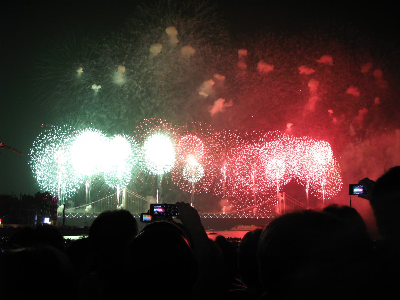 Fireworks bursting in green and red along the full span of the Gwangan Bridge in Busan, with cascades falling from the bridge deck into the water, seen over a dense crowd of silhouetted heads and raised cameras.