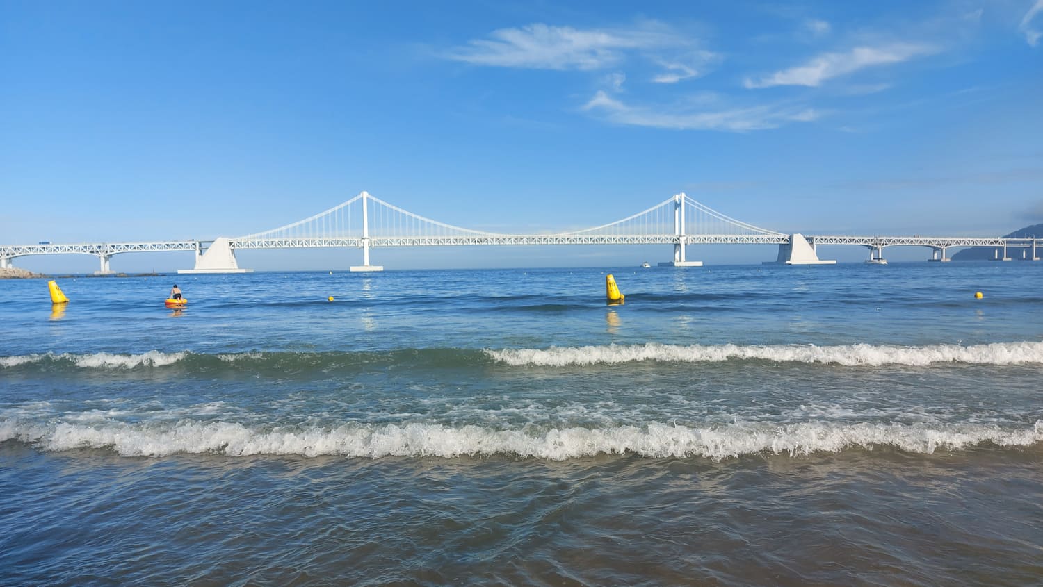 Gwangalli Beach in daylight, with waves in the foreground and the full white span of Gwangan Bridge directly ahead across the bay.