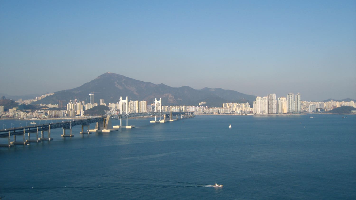 The full span of Gwangan Bridge seen from Igidae Park across the bay, with the Gwangalli and Haeundae skyline and a mountain ridge behind it.