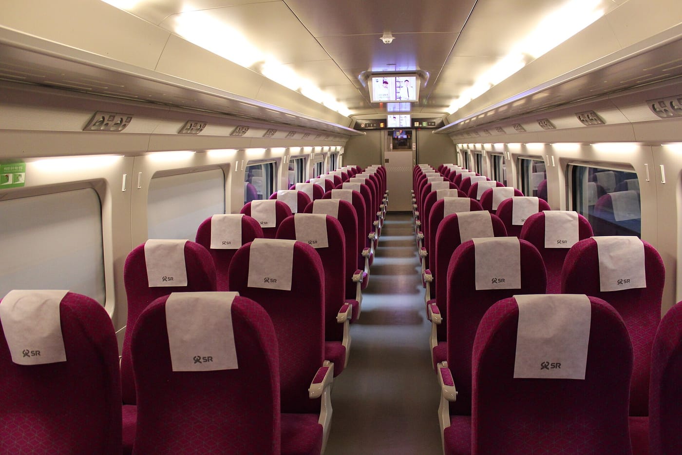 The interior of an SRT standard-class carriage, looking down the aisle. Rows of magenta seats with white headrest covers carrying the SR logo run to the end of the car, with an information screen above the far door.