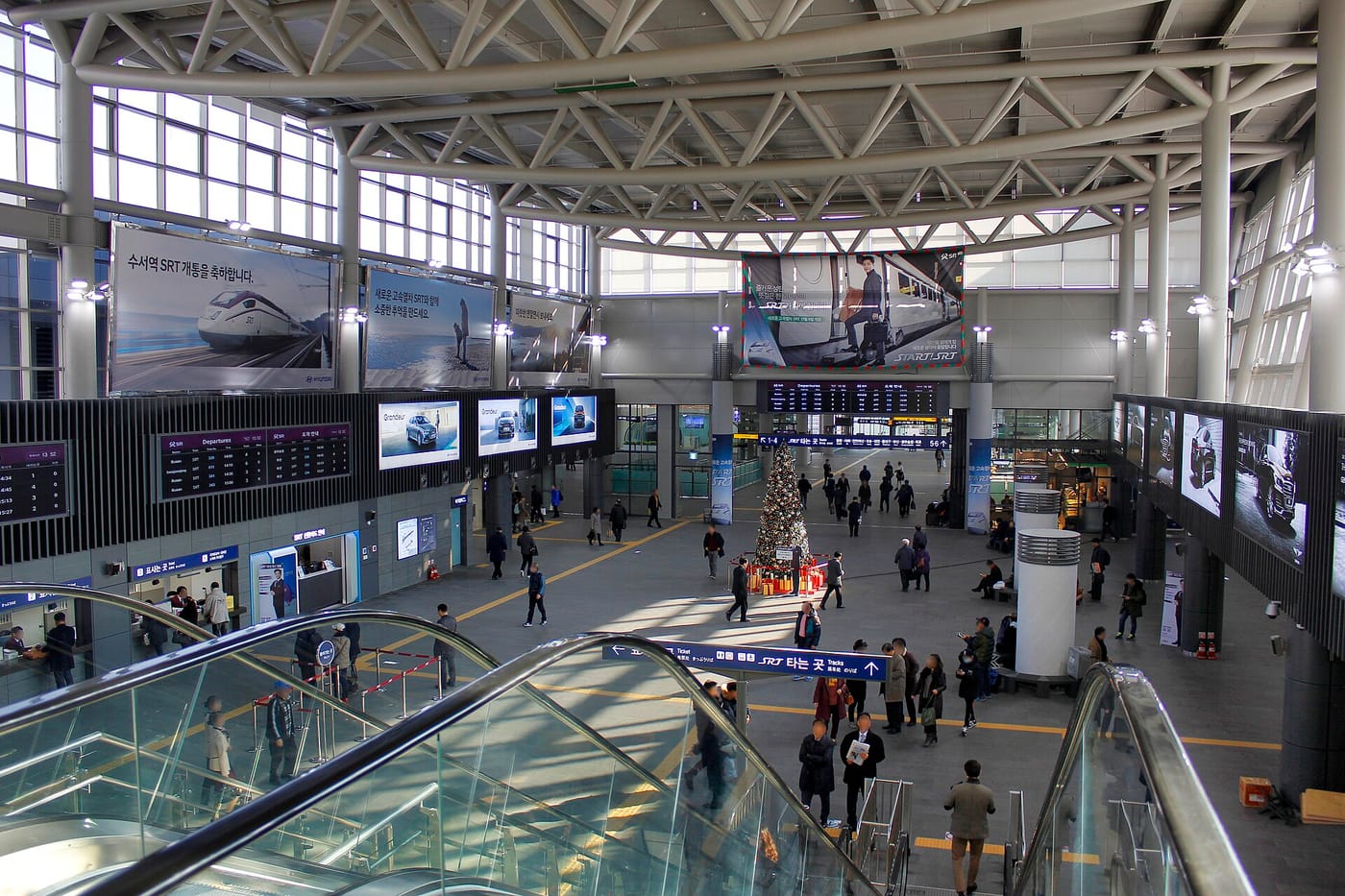The concourse of Suseo SRT station, seen from above an escalator. A large banner reads "수서역 SRT 개통을 축하합니다," departure boards and START SRT signage are visible, and travellers cross the polished floor below a steel-truss roof.