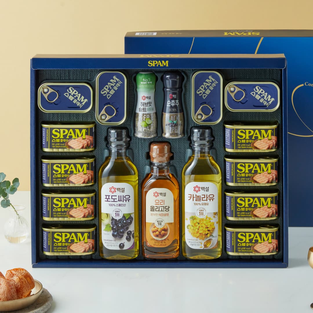 An open navy gift box containing eight 200g Spam cans and four small flat cans around the edges, with three bottles of Beksul grapeseed oil, cooking oligosaccharide syrup and canola oil in the centre, plus herb salt and black pepper grinders.