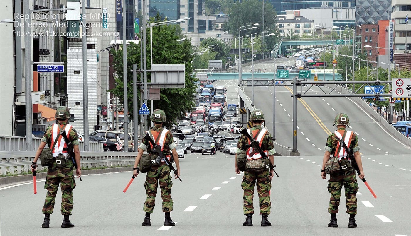 Four South Korean soldiers in helmets and white cross-belts standing in a line across an empty multi-lane road, holding red traffic batons, with stopped traffic visible in the distance behind them.