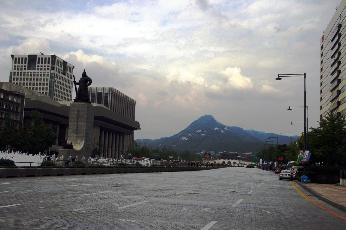 Sejong-daero seen from the south, a wide empty boulevard running north toward Gwanghwamun Gate and the mountain behind it, with the Admiral Yi Sun-sin statue on the left.