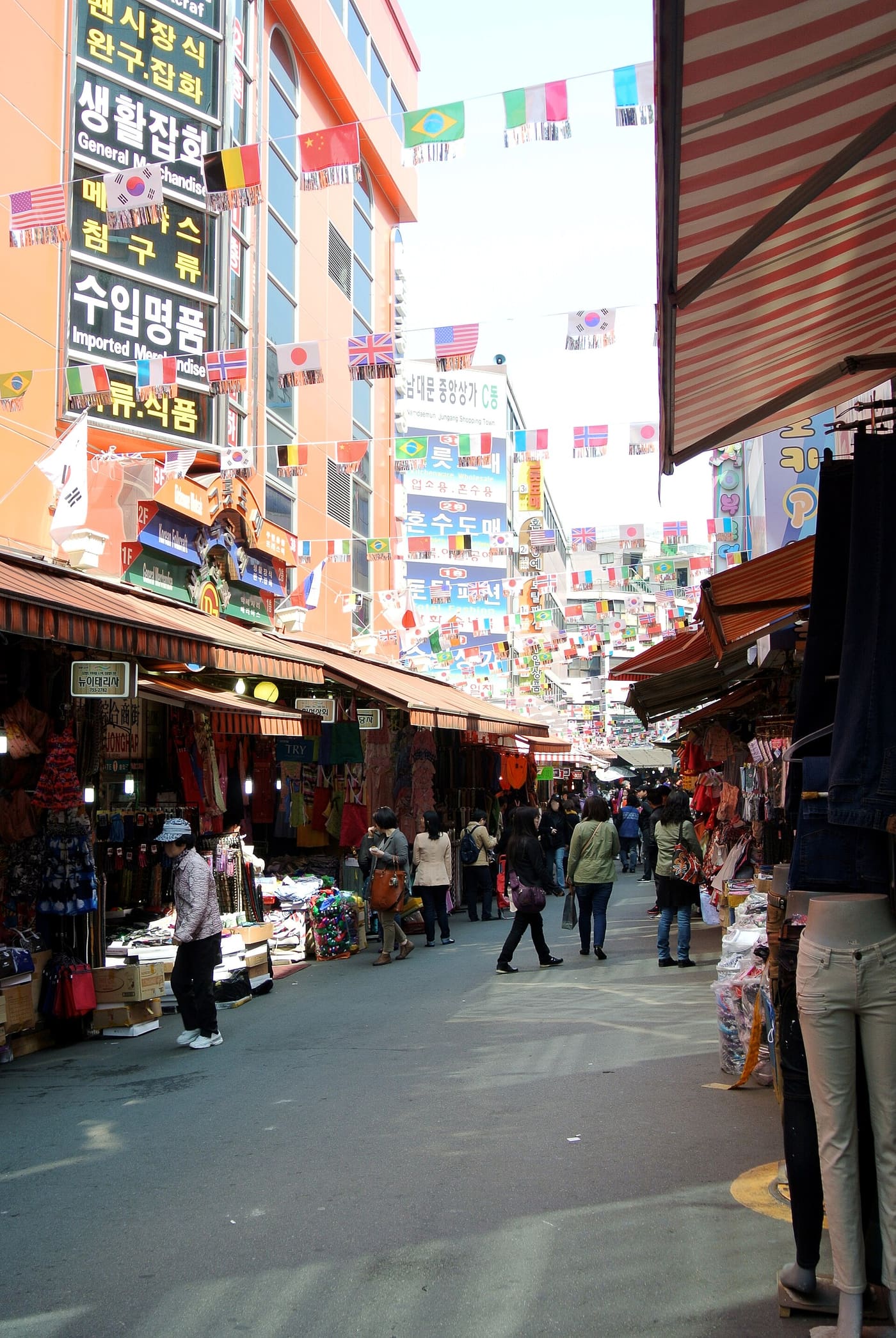 An alley inside Namdaemun Market with international flags strung overhead, a large sign reading Namdaemun Jungang Shopping Town C-dong, and shops selling bags and imported goods on both sides.