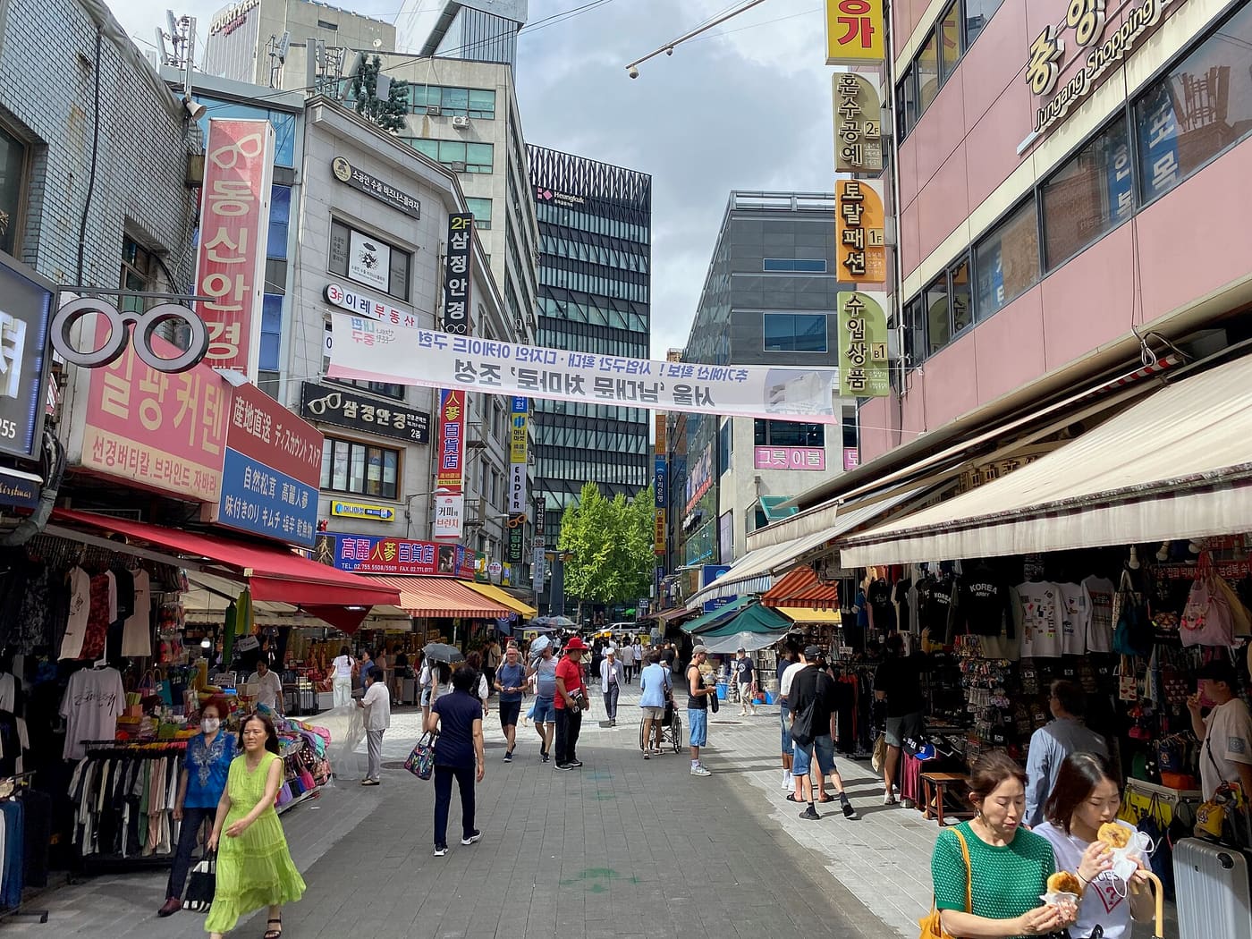 A busy Namdaemun Market street in Seoul lined with awnings and stalls, Korean shop signs on both sides, shoppers walking between racks of clothing and goods.